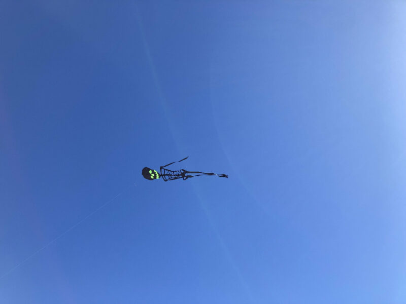 Photo of a black skeleton with neon green eye sockets and smile floating in a blue sky. One might or might not see a thin white line attached to it going down and out of view.